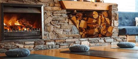 A stone fireplace with a fire, a stack of firewood, and yoga mats on the floor.