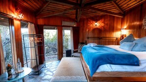 A wooden cabin bedroom with a bed, a small table with glasses, a chair, and a view of the outdoors.