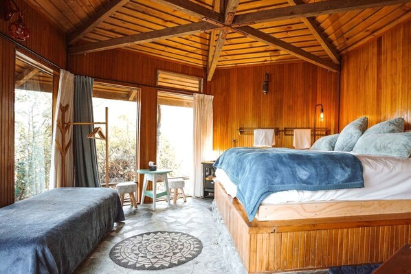 A wooden cabin bedroom with a bed, two bedside tables, a small table, and a window with curtains.