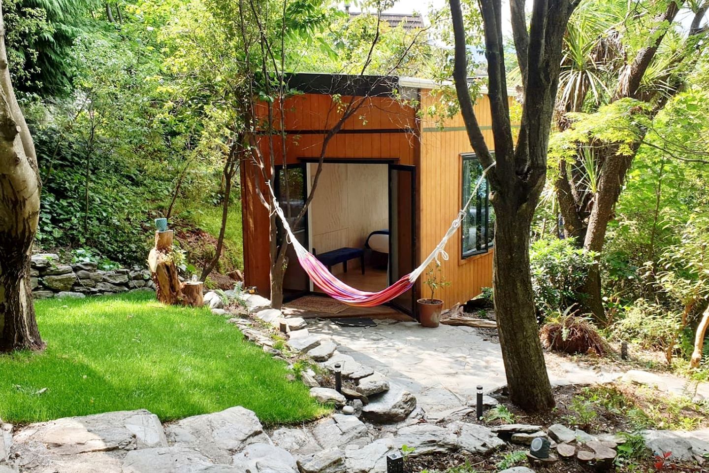 A wooden cabin with a hammock in front, surrounded by a stone pathway and lush greenery.