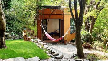 A wooden cabin with a hammock in front, surrounded by a stone pathway and lush greenery.