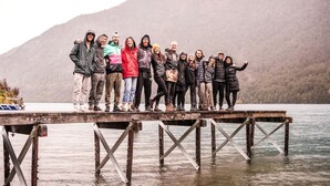 Eine Gruppe von Menschen steht auf einem hölzernen Steg, der sich über ein Gewässer erstreckt, mit Bergen im Hintergrund.