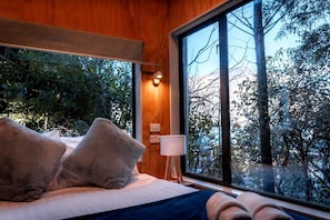 A bedroom with a large window, a bed with pillows, a bedside table, and a lamp.