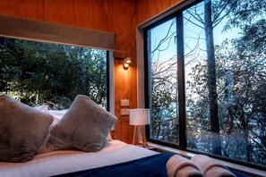 A bedroom with a large window, a bed with pillows, a bedside table, and a lamp.