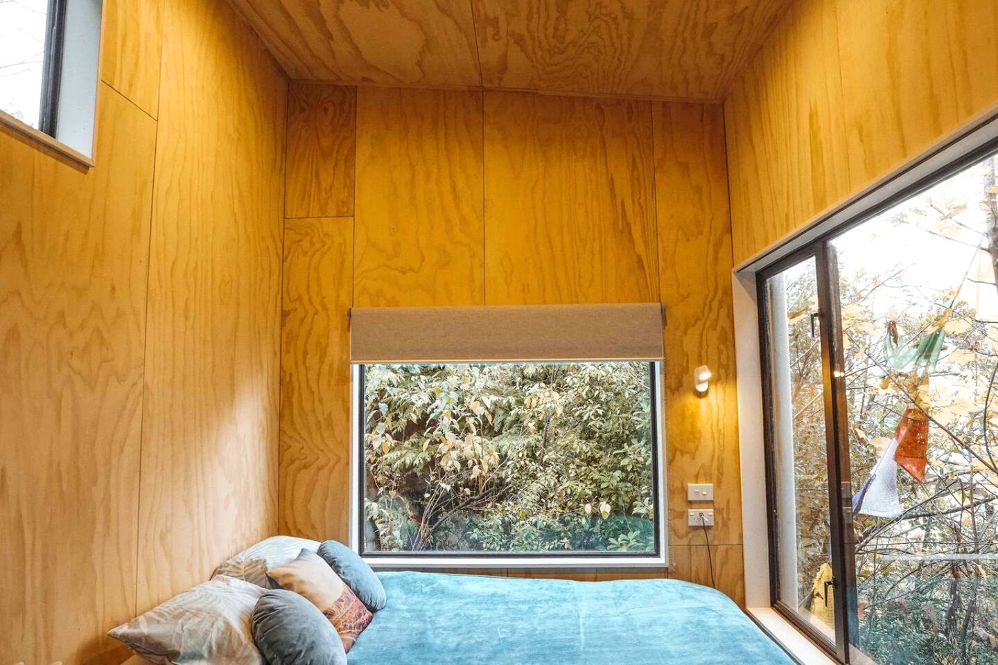 Une chambre avec des murs en bois, une grande fenêtre, un lit avec des oreillers et une vue sur les arbres.