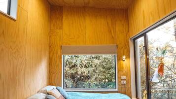 Une chambre avec des murs en bois, une grande fenêtre, un lit avec des oreillers et une vue sur les arbres.