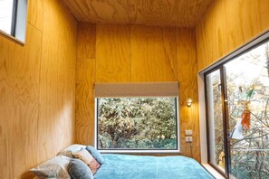 A bedroom with wooden walls, a large window, a bed with pillows, and a view of trees.