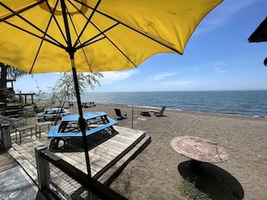 A beachside patio with wooden tables and chairs, a yellow umbrella, and a view of the ocean.