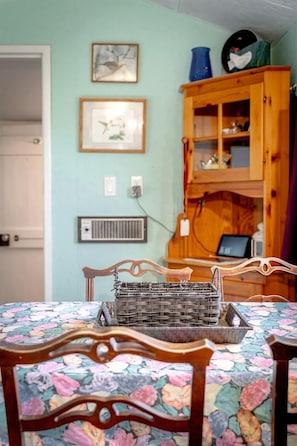 A dining area with a floral tablecloth, wooden chairs, a wicker basket, a wooden cabinet with glass doors, and wall-mounted electrical outlets. - Cozy Lake Tiny House Incredible to Create New Memories in Ontario (Leamington)