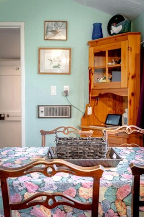 Un coin repas avec une nappe à motifs floraux, des chaises en bois, un panier en osier, un buffet en bois doté de portes en verre et des prises électriques fixées au mur.