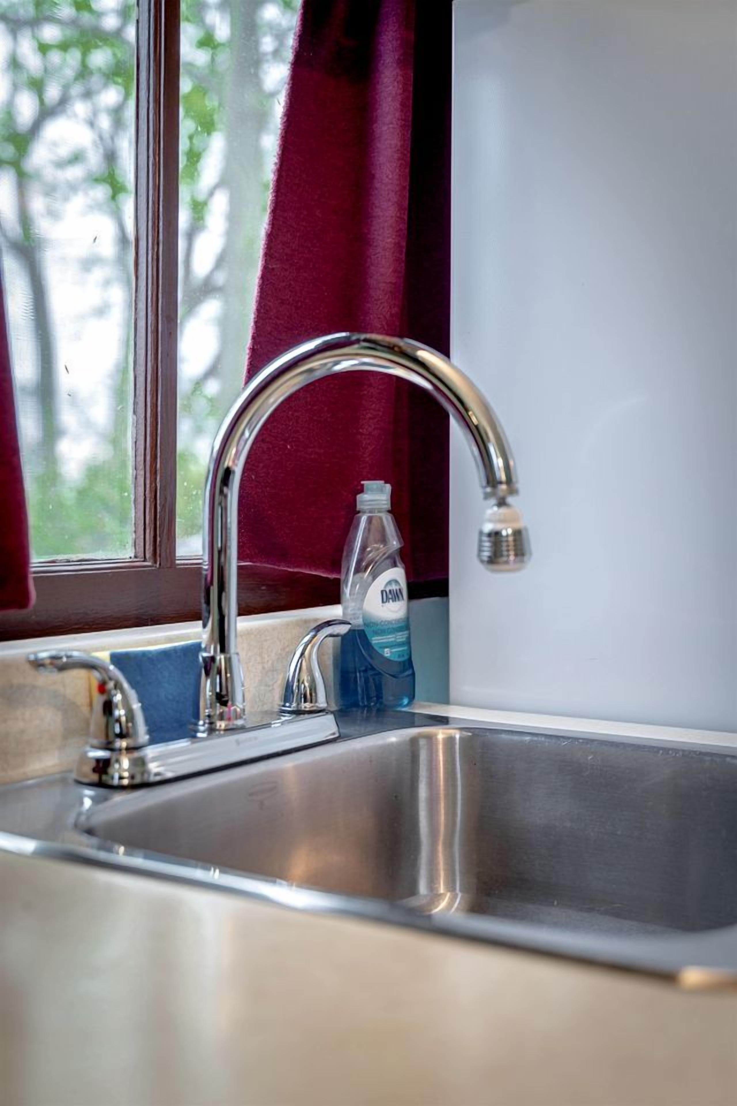 A stainless steel sink with a single faucet, a bottle of hand soap, and a window with curtains.