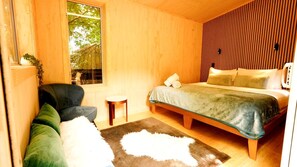 Ein Zimmer aus Holz mit einem Bett, einem Sessel, einem kleinen Tisch und einem Fenster.