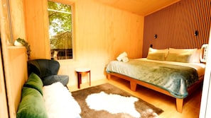 A wooden room with a bed, a chair, a small table, and a window.