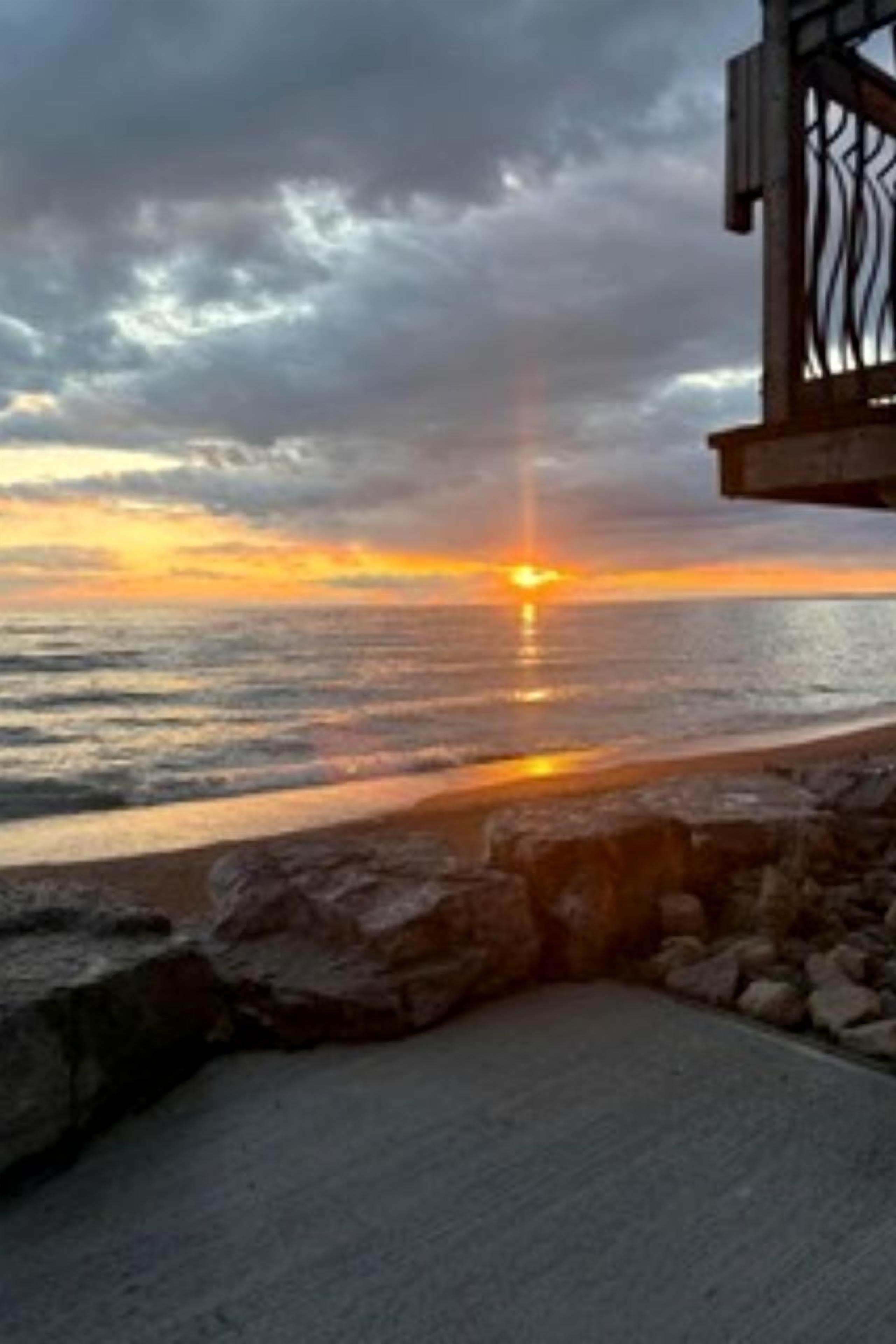 A sunset over the ocean with a rocky shoreline and a structure with metal railings.