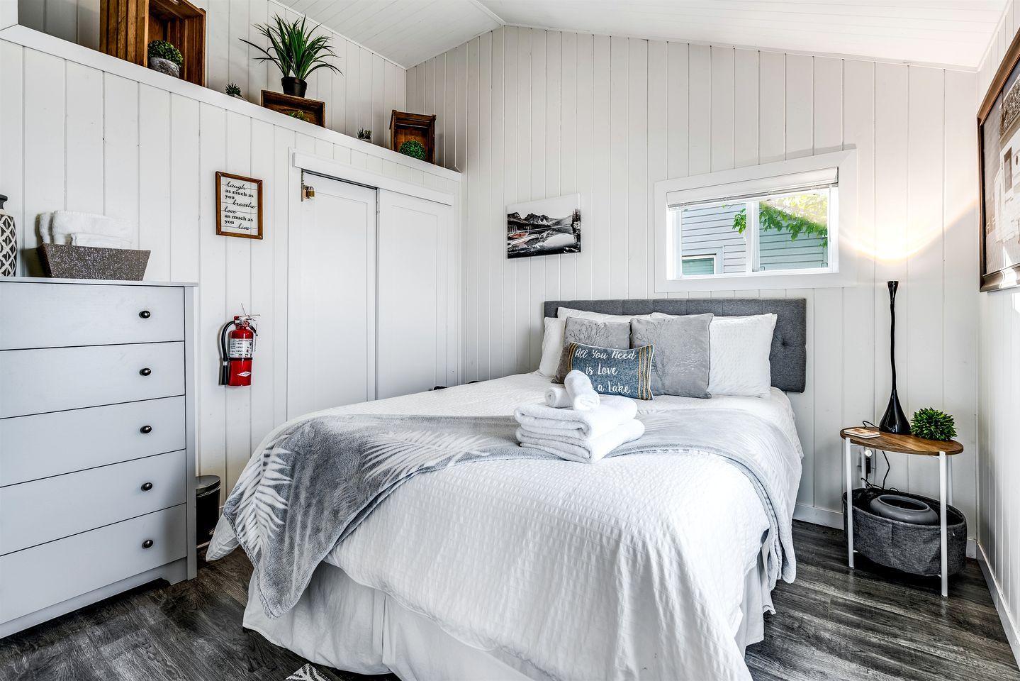 Een slaapkamer met een bed, een dressoir, een brandblusser, een raam met uitzicht en een nachtkastje met een lamp.