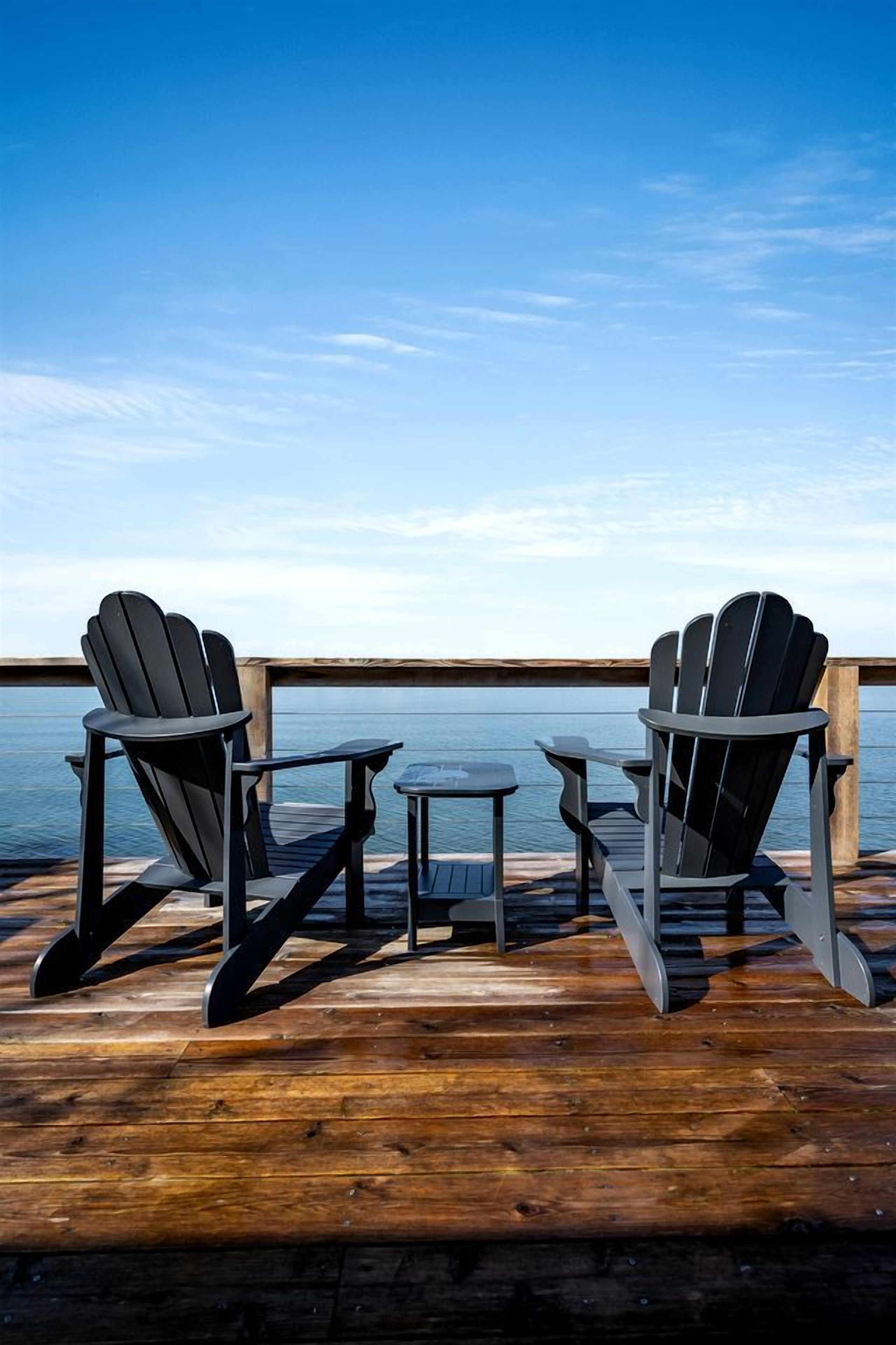 Two Adirondack chairs and a small table on a wooden deck overlooking a body of water.
