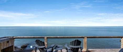 Een houten terras met drie Adirondack-stoelen gericht op een rustige zee onder een heldere lucht.