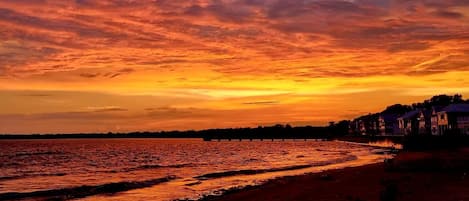 A beach with a sunset, a pier, and houses along the shore.