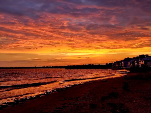 A beach with a sunset, a pier, and houses along the shore.