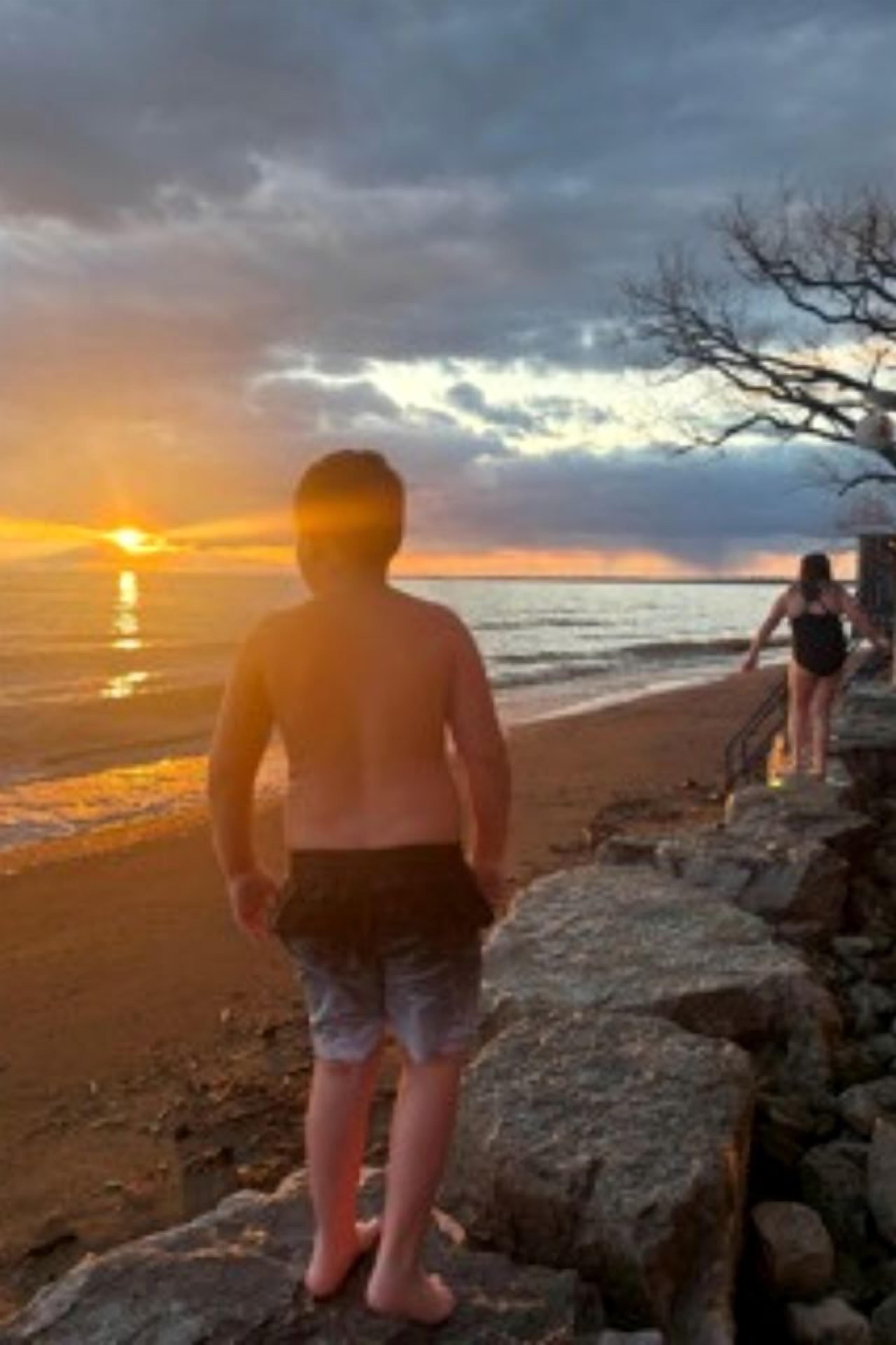 Escena de playa con atardecer, costa rocosa y una persona caminando.