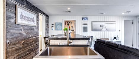 A modern kitchen with a large stainless steel sink, wooden wall paneling, a dining area with a dark-colored sofa, and a view of the beach.
