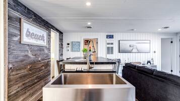 A modern kitchen with a large stainless steel sink, wooden wall paneling, a dining area with a dark-colored sofa, and a view of the beach.