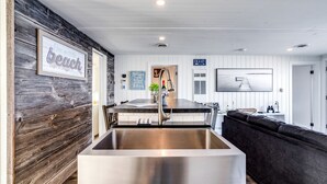 Una cocina moderna con un lavamanos grande de acero inoxidable, paneles de madera en la pared, un área de comedor con un sofá de color oscuro y vistas a la playa.