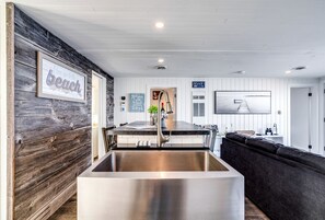 A modern kitchen with a large stainless steel sink, wooden wall paneling, a dining area with a dark-colored sofa, and a view of the beach. - Sweet Beach Rental with Jacuzzi, Deck and Amazing Sunsets in Ontario (Leamington)