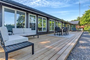 A modern building with large windows, a wooden deck, outdoor seating, and a small table. - Charming Cottage with Indoor Heated Pool near Sandbanks Provincial Park in Picton, Ontario (Prince Edward)