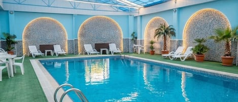 An indoor swimming pool with a glass ceiling, surrounded by lounge chairs and potted plants.