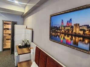 Habitación de hotel con cama, mesita de noche con tetera y tazas, un cuadro en el muro con una vista urbana, y vista al horizonte de la ciudad.