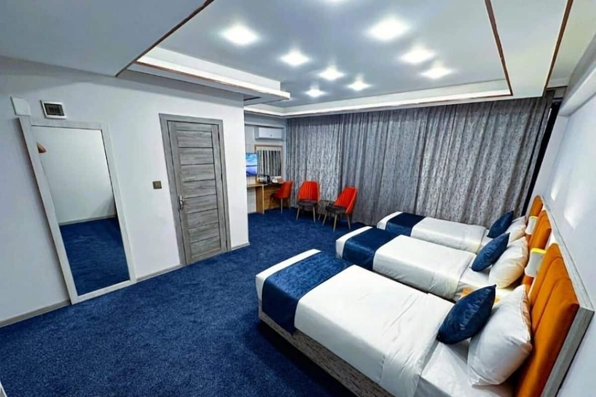 Habitación de hotel con alfombra azul, dos camas con ropa de cama blanca y azul, una cortina gris y una pequeña mesa con sillas rojas.
