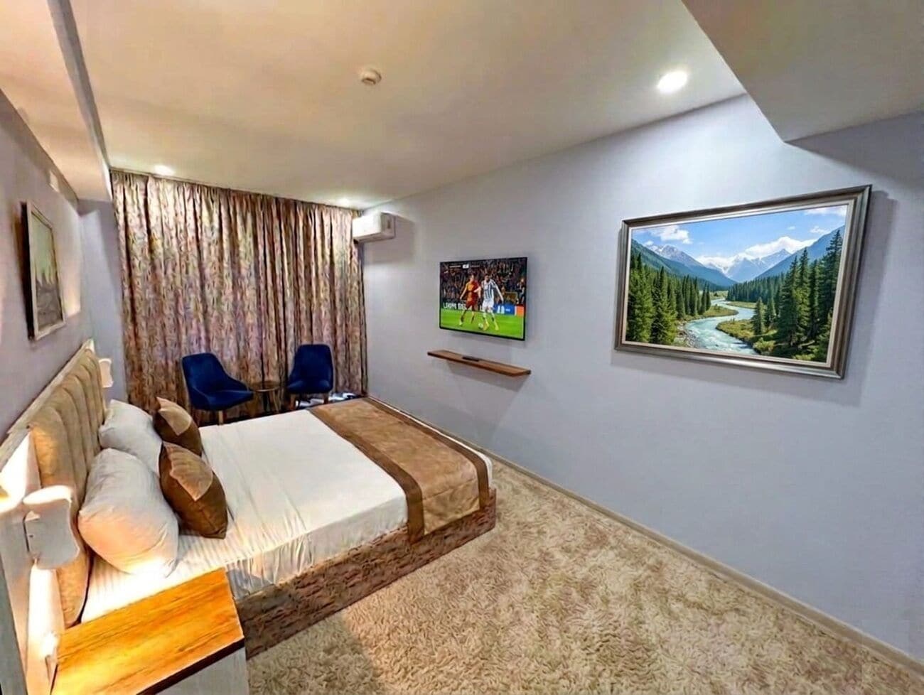 A hotel room with a bed, a TV, a painting, and a view of a mountainous landscape.