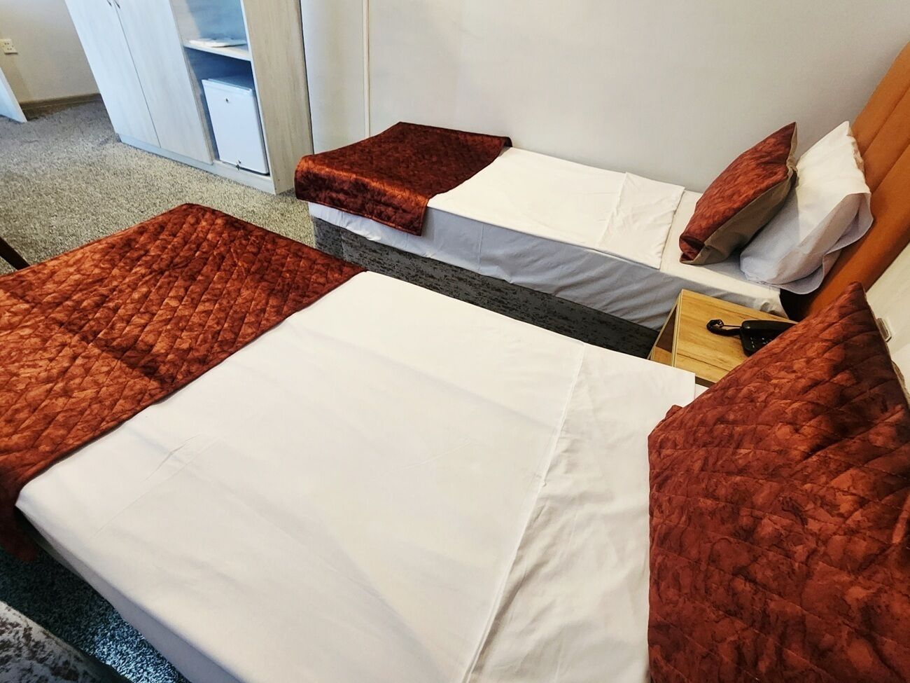 Une chambre d’hôtel avec deux lits, chacun recouvert d’une couette rouge et de draps blancs. Une table de chevet en bois avec une lampe se trouve entre les deux lits.