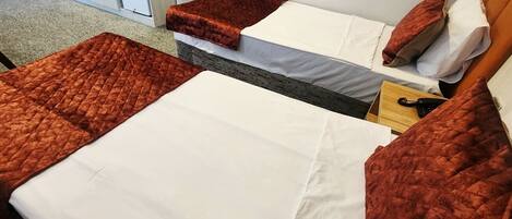 Une chambre d’hôtel avec deux lits, chacun recouvert d’une couette rouge et de draps blancs. Une table de chevet en bois avec une lampe se trouve entre les deux lits.