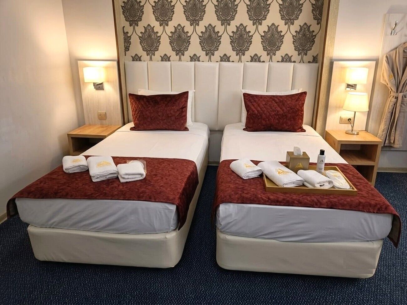 Habitación de hotel con dos camas, cada una con cubrecama rojo, ropa blanca y toallas. Mesitas de noche con lámparas y un neceser.