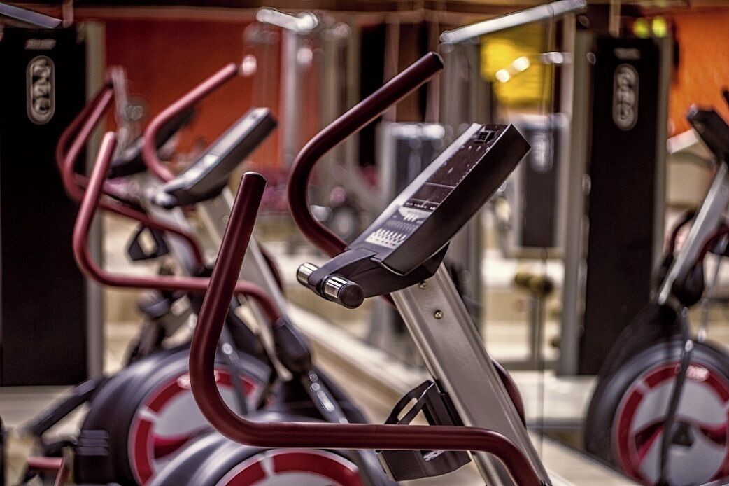 A row of stationary bikes in a gym with a mirrored wall and a door in the background.