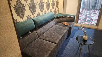 A room with a sectional sofa, a small round table with a vase, patterned wallpaper, and a patterned carpet.