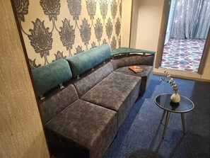 Ein Zimmer mit einer Eckcouch, einem kleinen runden Tisch mit einer Vase, strukturierten Tapeten und einem gemusterten Teppich.