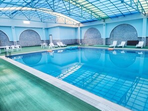 Una piscina interior con agua azul cristalina, rodeada de sillas reclinables blancas y una pared de azulejos.