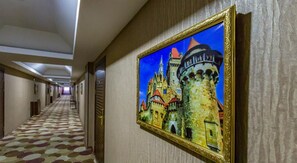 A hotel hallway with a patterned carpet, a framed painting of a castle, and doors along the corridor. - Amazon Hotel - Full Board, Breakfast, Lunch & Dinner Included (Baku)