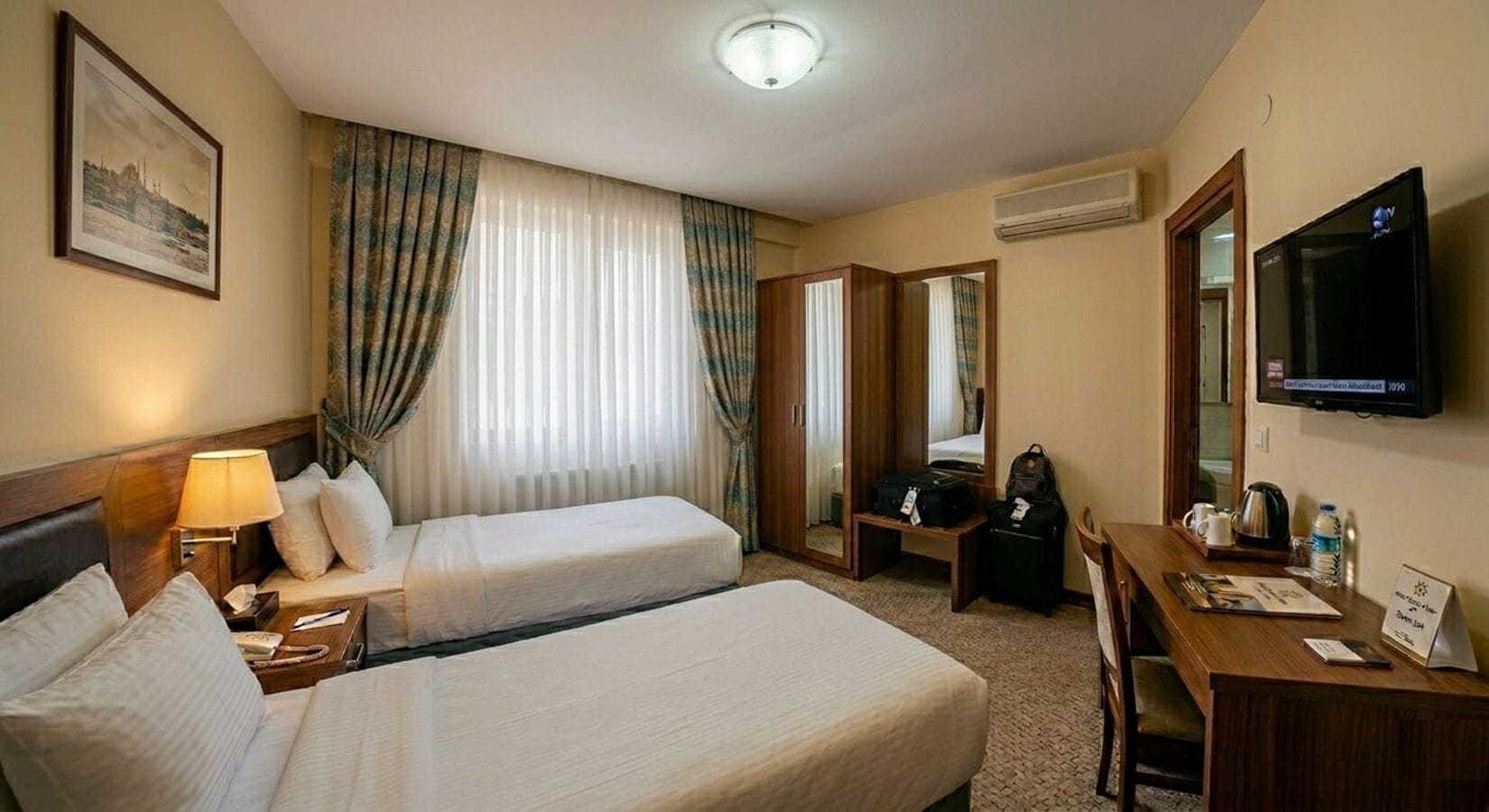 Ein Hotelzimmer mit zwei Betten, einem Schreibtisch, einem Fernseher und einem Fenster mit Vorhängen.