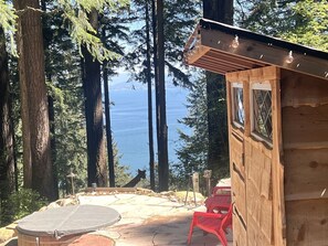 Une cabane en bois avec une terrasse en pierre, une chaise rouge et une vue sur la forêt et l’eau.