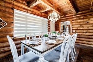 Une salle à manger aménagée dans une cabane en rondins, avec une table en bois dressée pour quatre personnes, des chaises blanches, un lustre et une grande fenêtre dotée de stores.