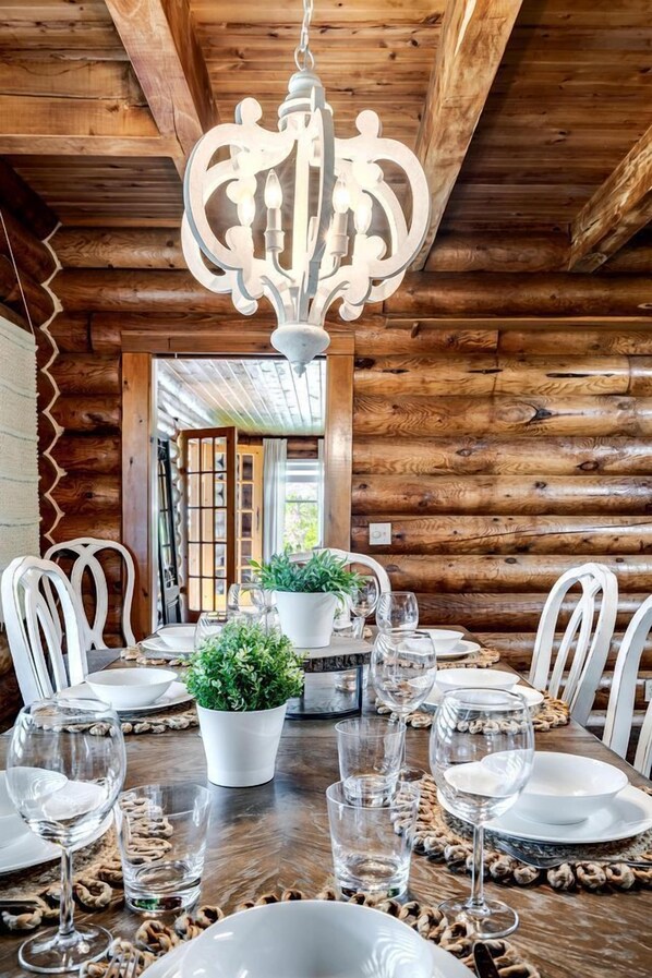 L’intérieur d’un chalet en bois, avec une table à manger dressée pour six personnes, des chaises blanches, un lustre et des plantes en pot.