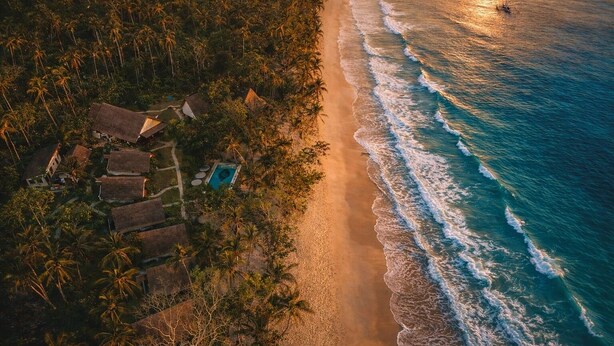 Vista aérea de uma praia com palafitas de telhado de palha e piscina, cercada por densas palmeiras.
