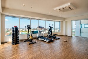 A spacious gym with a treadmill, elliptical, and weight rack, large windows with a city view. - Grand Kamala Lagoon Meeran By Reccoma (Bekasi)