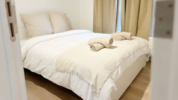 Una cama bien hecha con edredón blanco y manta color arena, piso de madera y cortinas color beige.