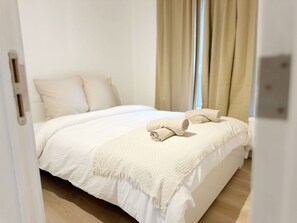 Una cama bien hecha con edredón blanco y manta color arena, piso de madera y cortinas color beige.
