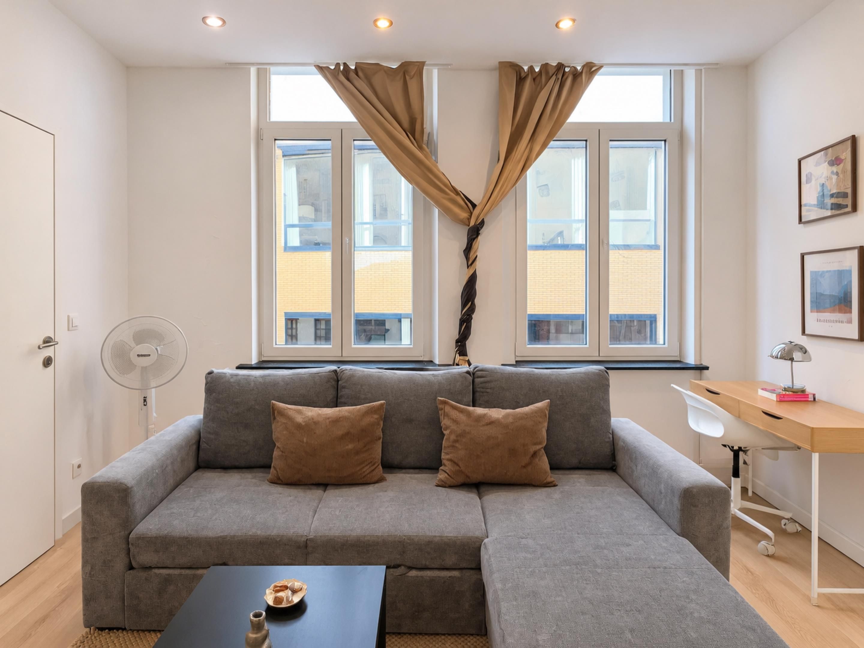 Een moderne woonkamer met een grijze bank, twee kussens, een bijzettafeltje met een vaas en een schaal, een ventilator en een bureau met een lamp en boeken.
